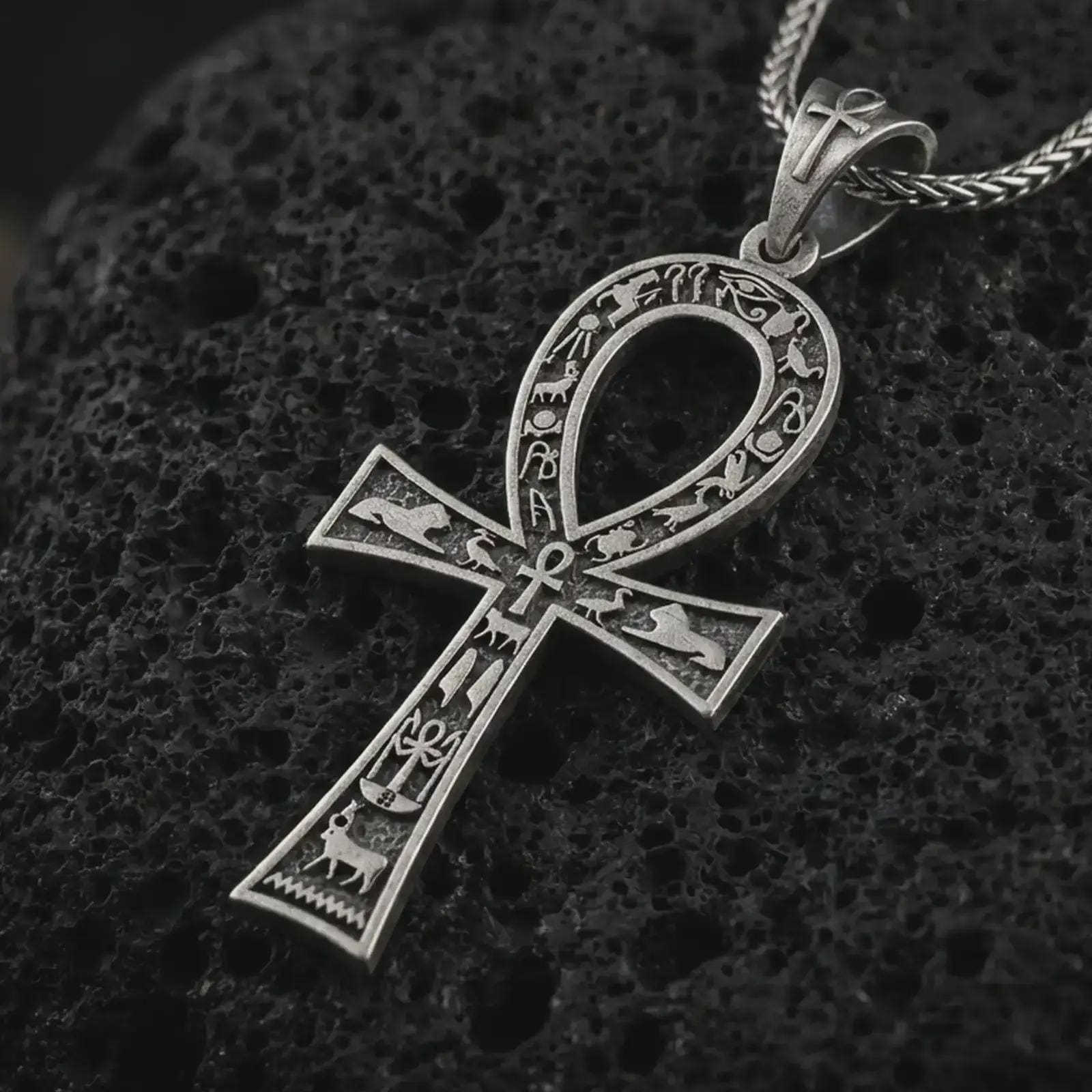 Oxidized ankh cross pendant necklace on textured volcanic rock, close-up studio shot highlighting the ancient Egyptian key of life symbol, hieroglyphic details, and antique silver contrast.