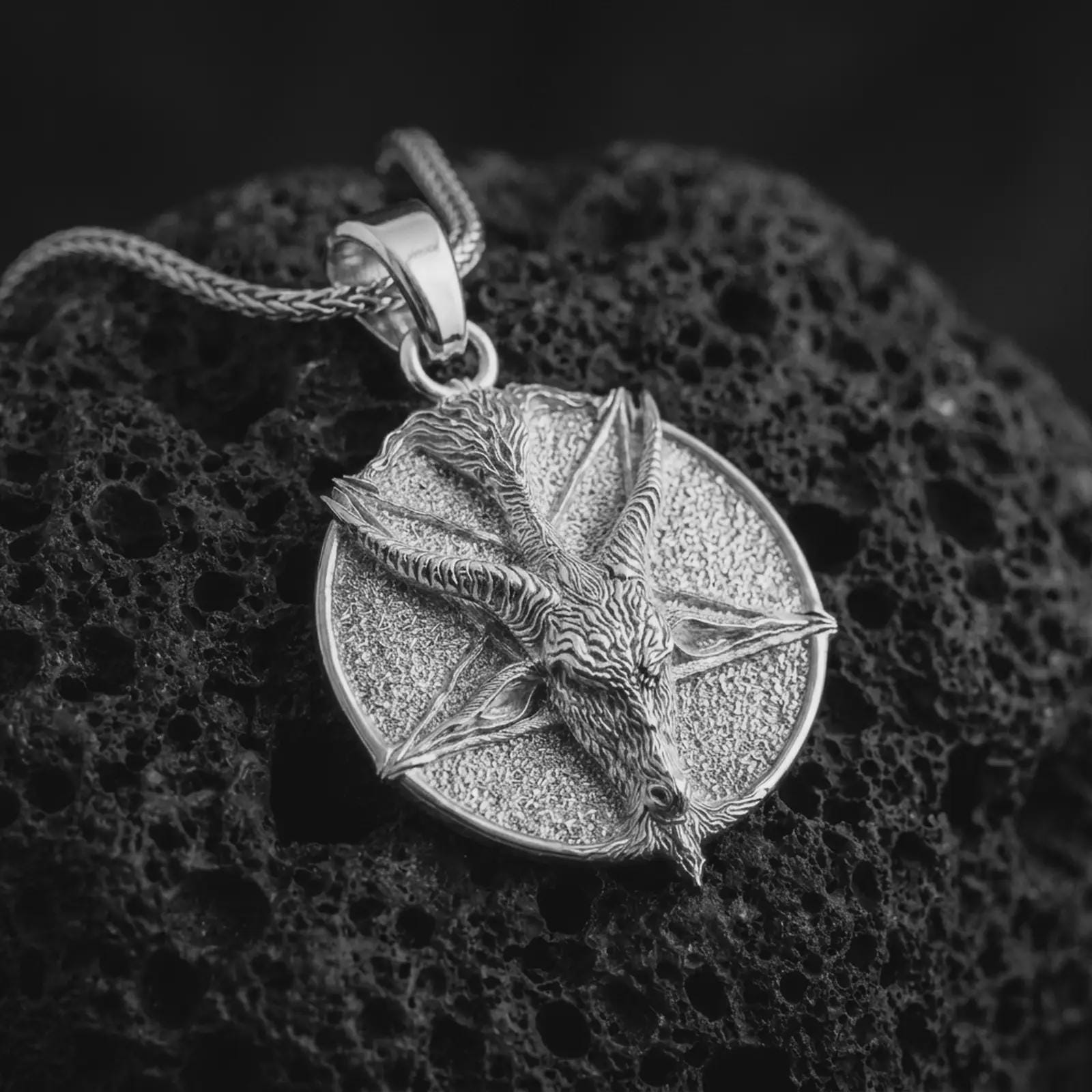 Polished silver goat pentagram pendant necklace on textured volcanic rock, centered macro jewelry photo with detailed horned goat relief and bright silver highlights.