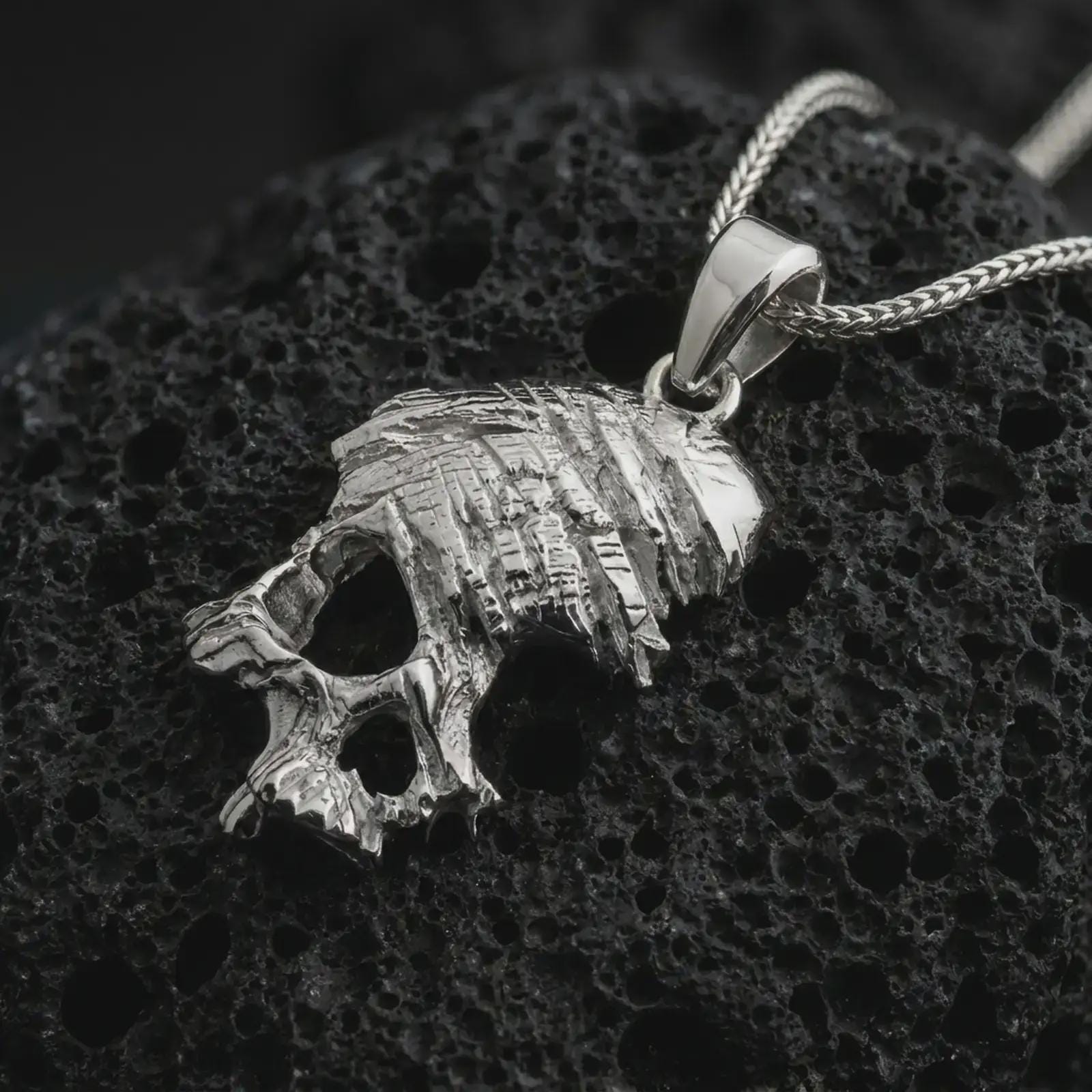 Polished silver cracked skull pendant necklace on textured volcanic rock, centered macro jewelry photo with detailed bone structure, broken surface effect, and bright silver highlights.