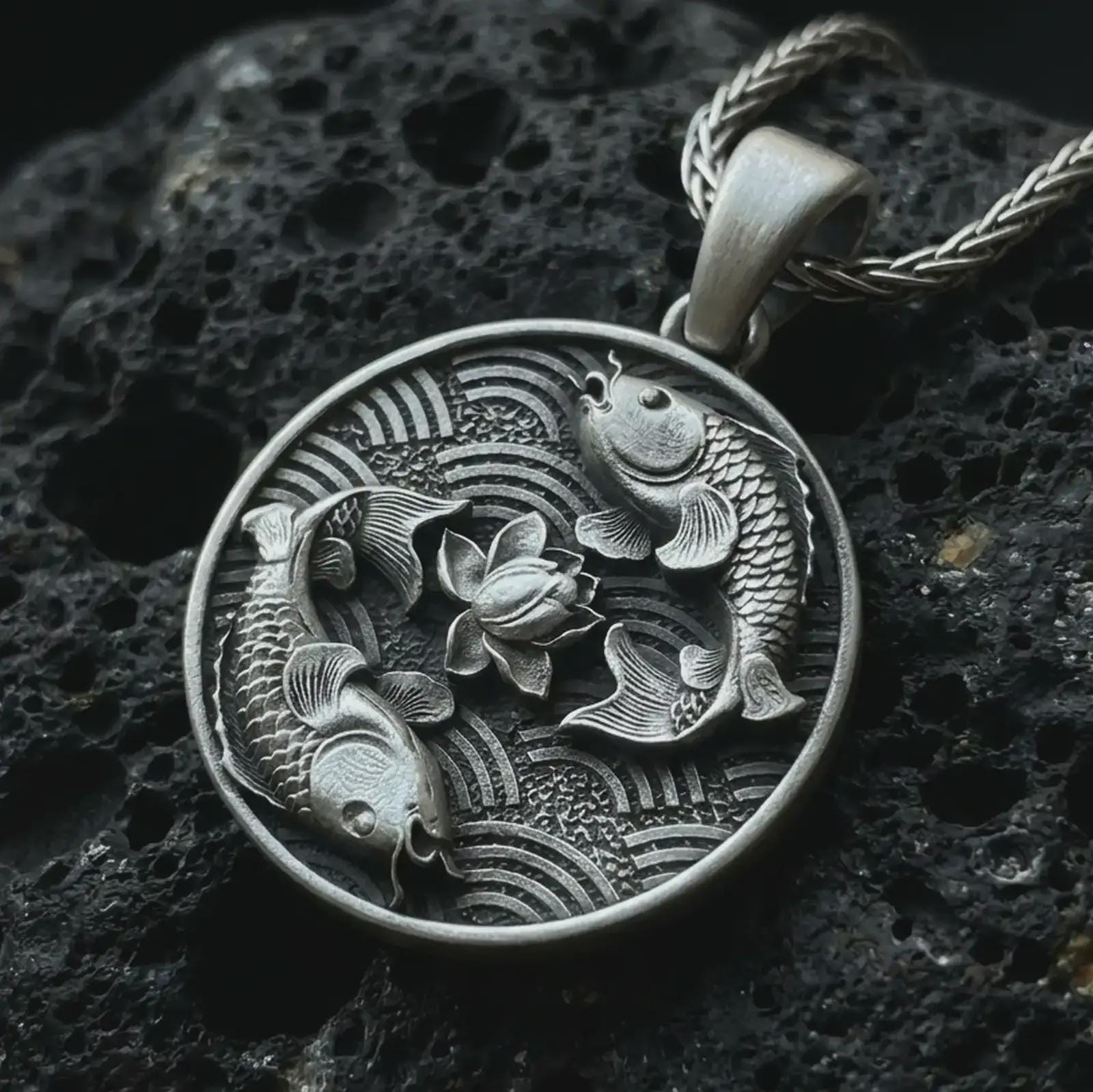 Oxidized koi fish lotus pendant necklace on textured volcanic rock, close-up studio shot highlighting the twin koi relief, lotus flower center, wave pattern, and antique silver contrast.