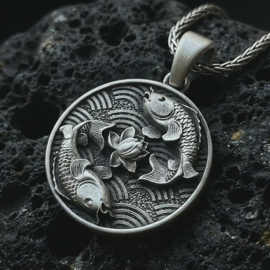 Oxidized koi fish lotus pendant necklace on textured volcanic rock, close-up studio shot highlighting the twin koi relief, lotus flower center, wave pattern, and antique silver contrast.
