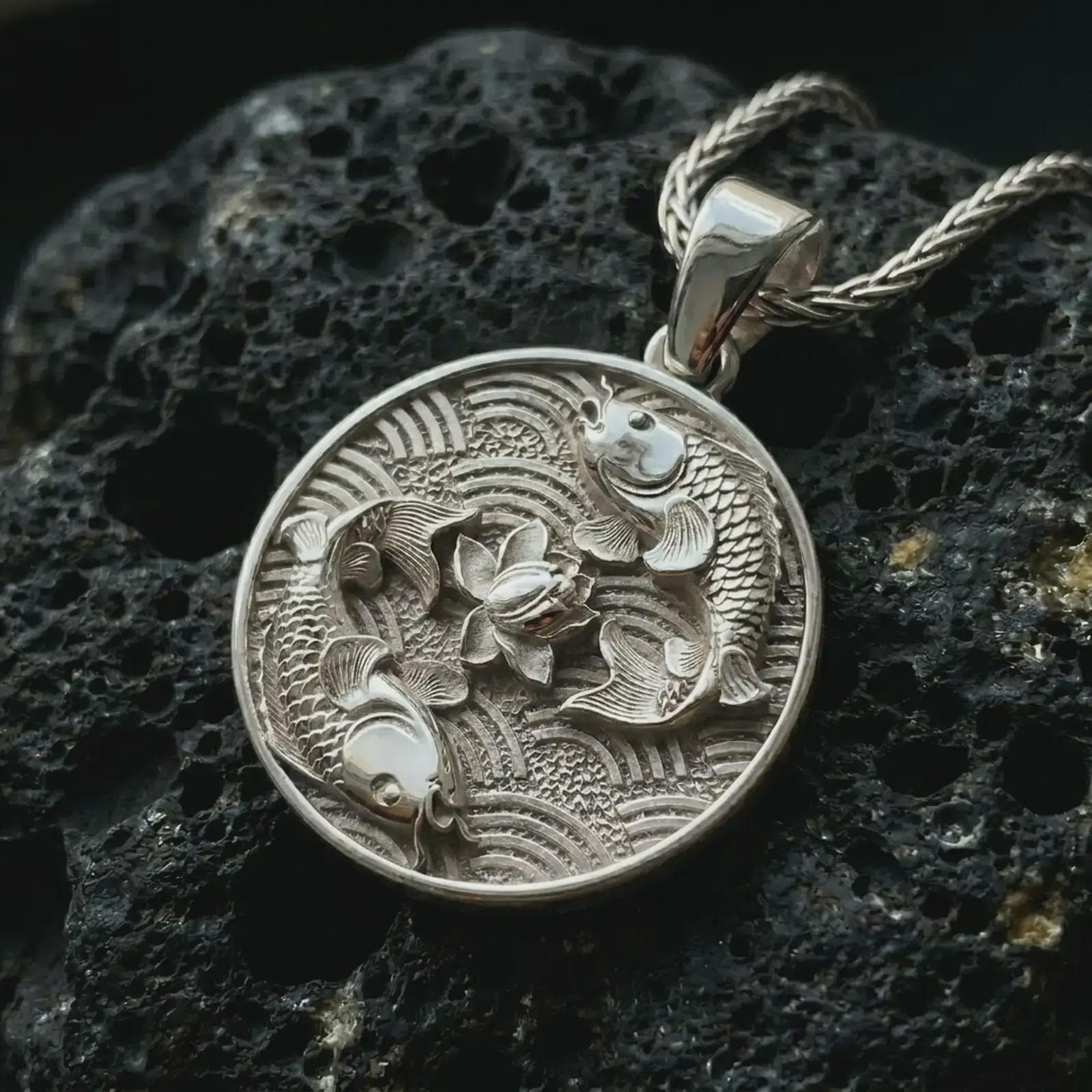 Polished silver koi fish lotus pendant necklace on textured volcanic rock, centered macro jewelry photo with detailed fish scales, flowing fins, and bright silver highlights.