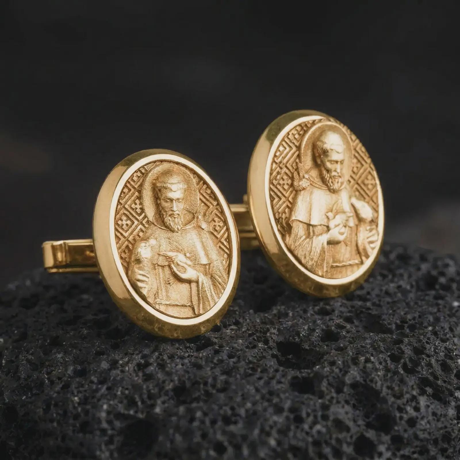 Gold plated Saint Francis cufflinks sit on textured volcanic rock in a dramatic studio macro shot, highlighting the sacred saint medallion design.