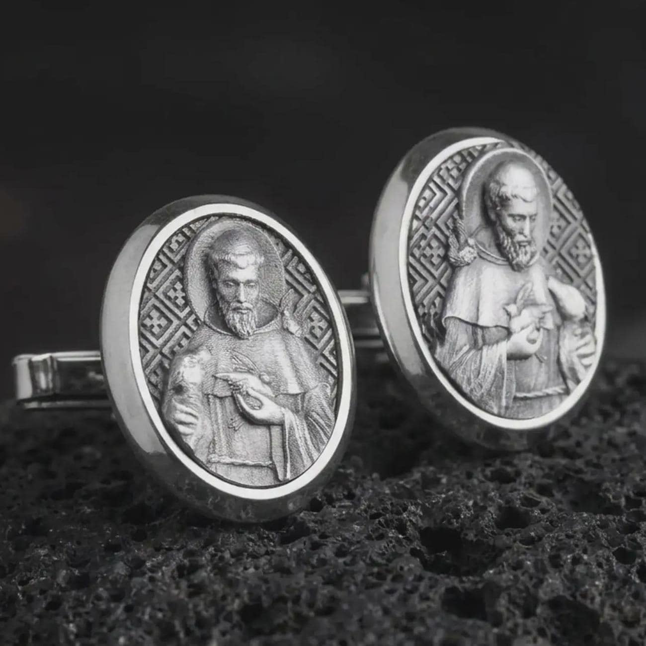 An oxidized Saint Francis cufflink pair rests on dark volcanic rock in a macro studio close-up, showing a detailed Catholic saint relief inside oval frames.