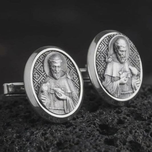 An oxidized Saint Francis cufflink pair rests on dark volcanic rock in a macro studio close-up, showing a detailed Catholic saint relief inside oval frames.