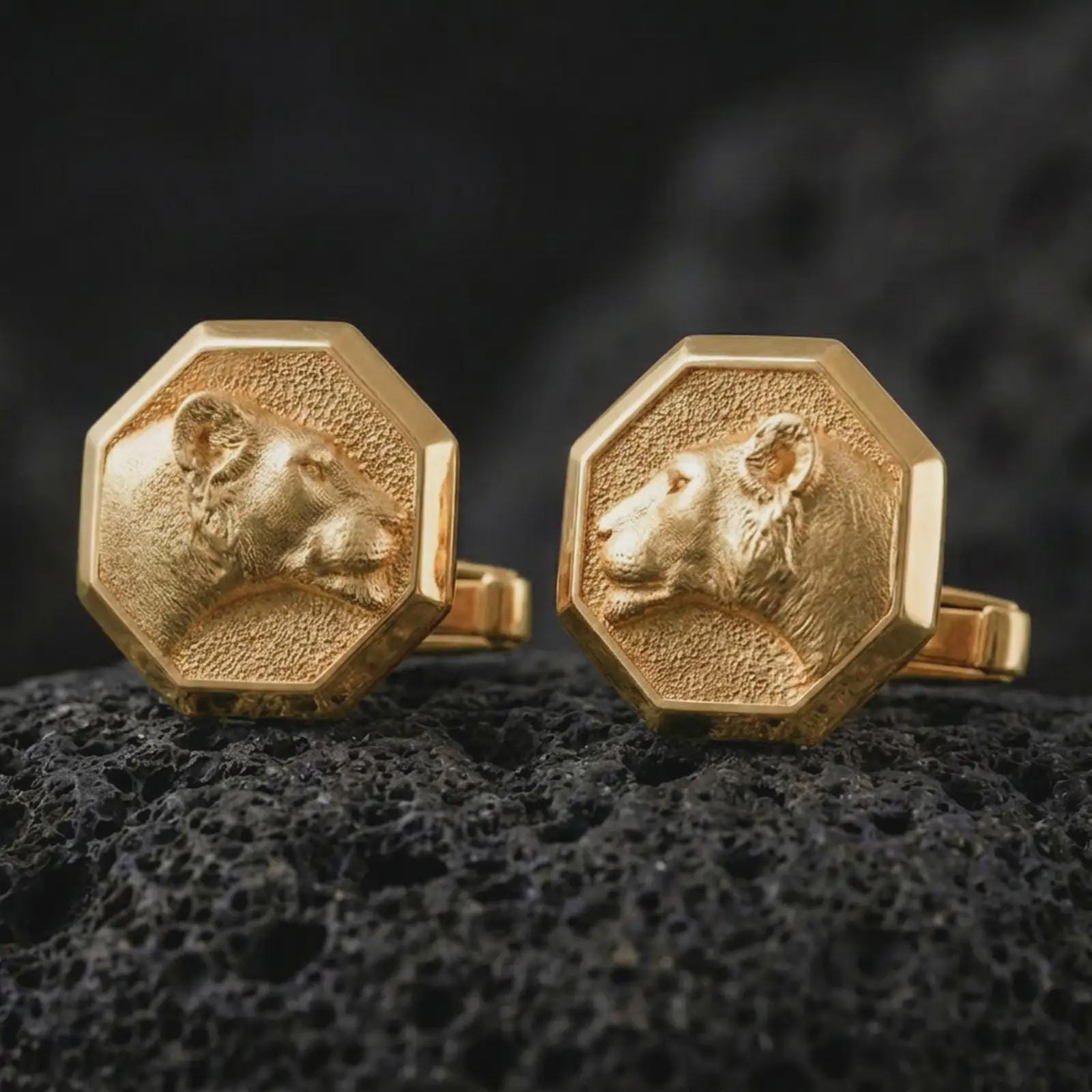Gold plated lioness head cufflinks sit on textured volcanic rock in a dramatic studio macro shot, highlighting the elegant animal motif and formal accessory design.