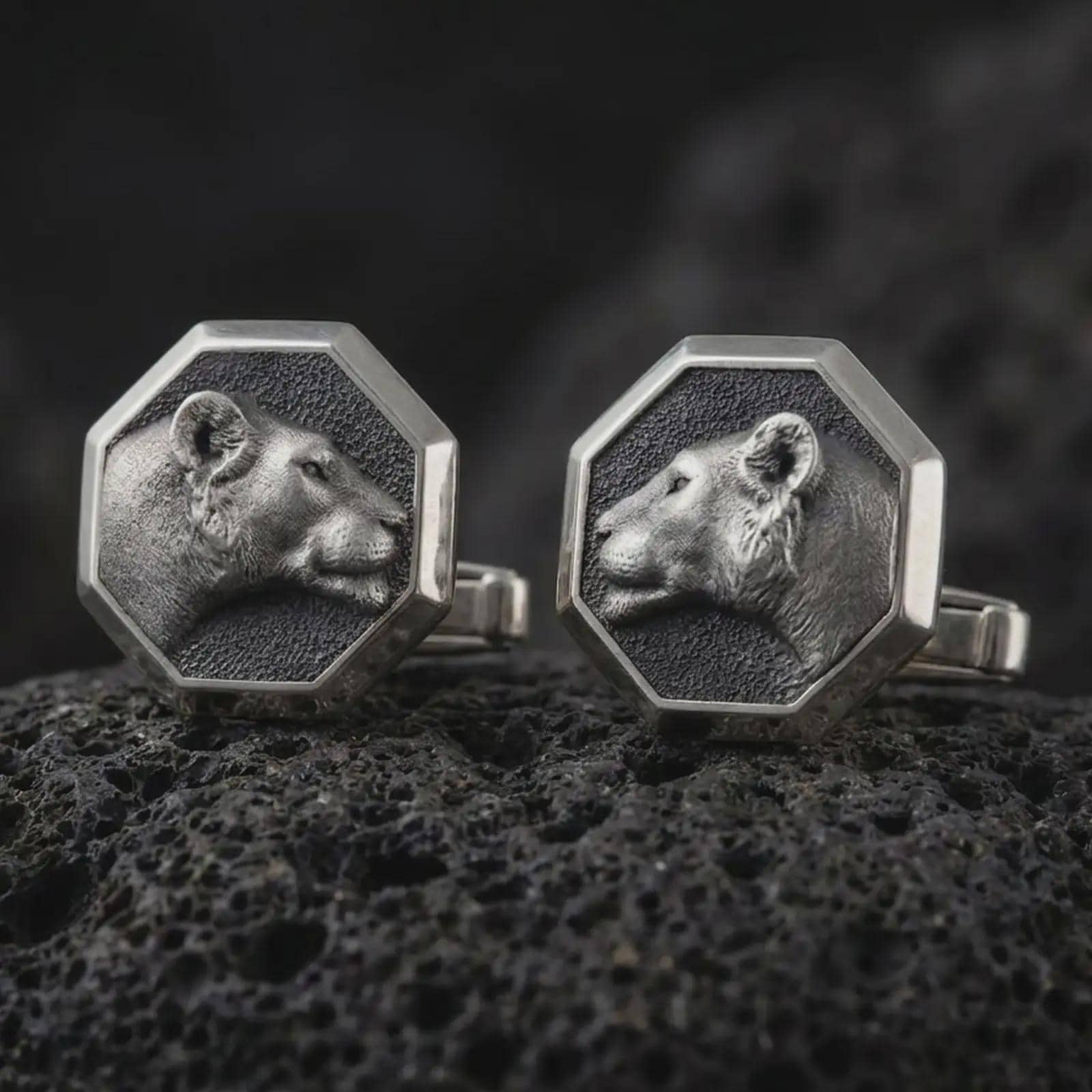 An oxidized lioness head cufflink pair rests on dark volcanic rock in a macro studio close-up, showing a sculpted wild cat profile inside octagon frames.
