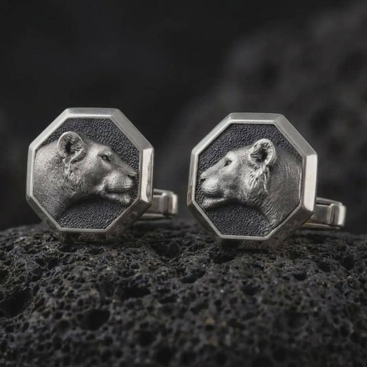 An oxidized lioness head cufflink pair rests on dark volcanic rock in a macro studio close-up, showing a sculpted wild cat profile inside octagon frames.