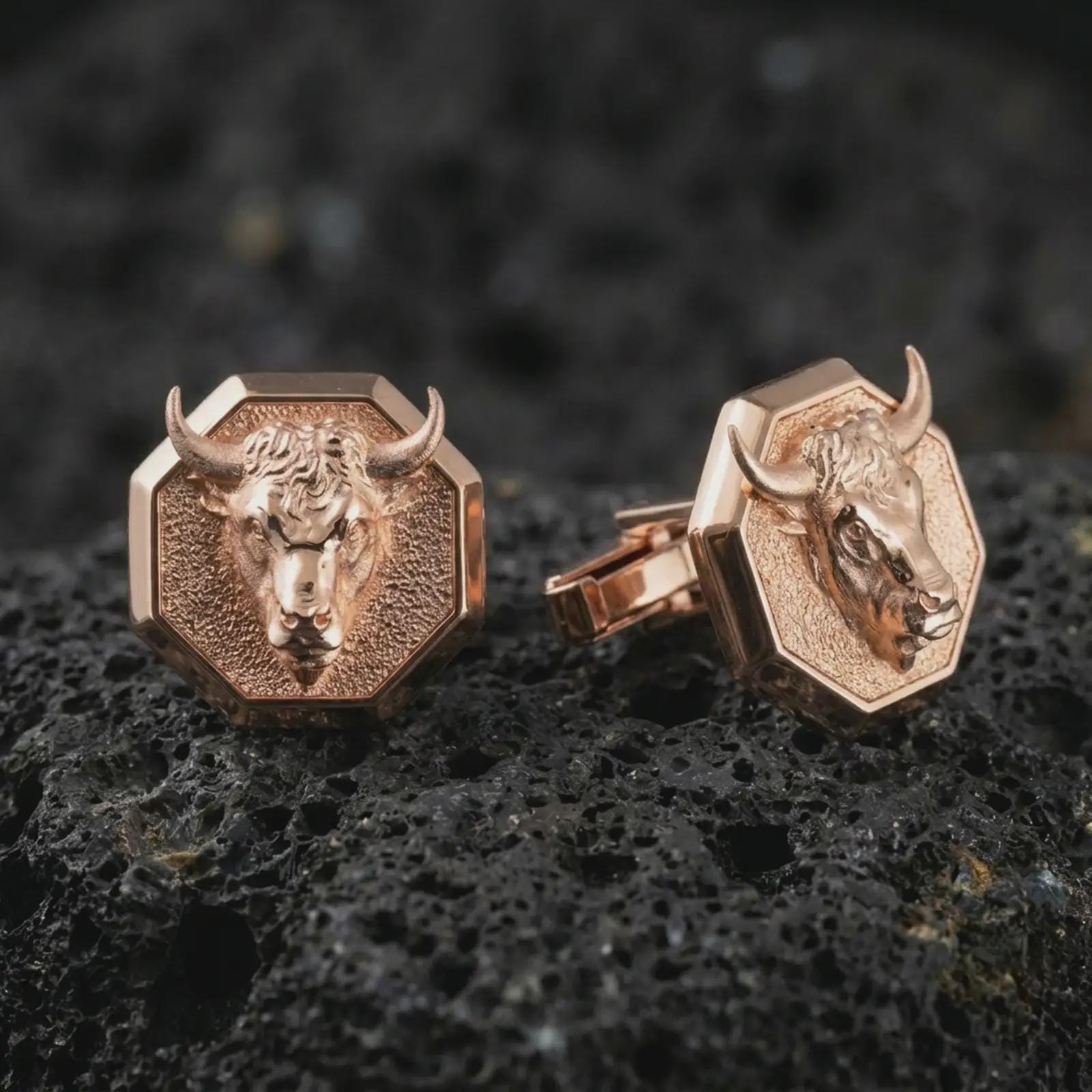 Rose gold plated bull head cufflinks are photographed on porous volcanic rock in a close-up studio composition with refined luxury lighting and strong bull features.