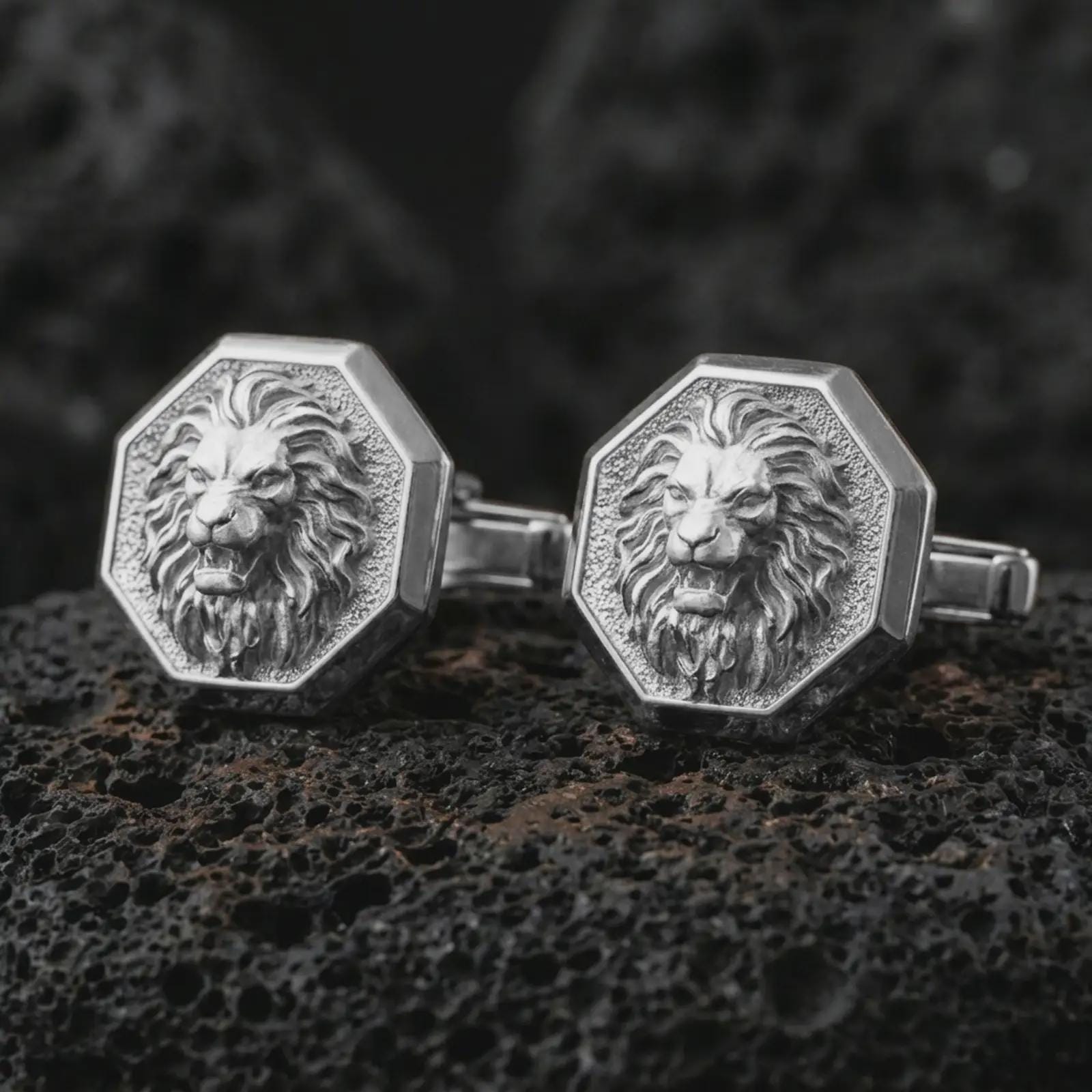 Polished silver lion head cufflinks are displayed on black volcanic rock in a premium macro studio close-up with sharp facial texture and clean geometric edges.