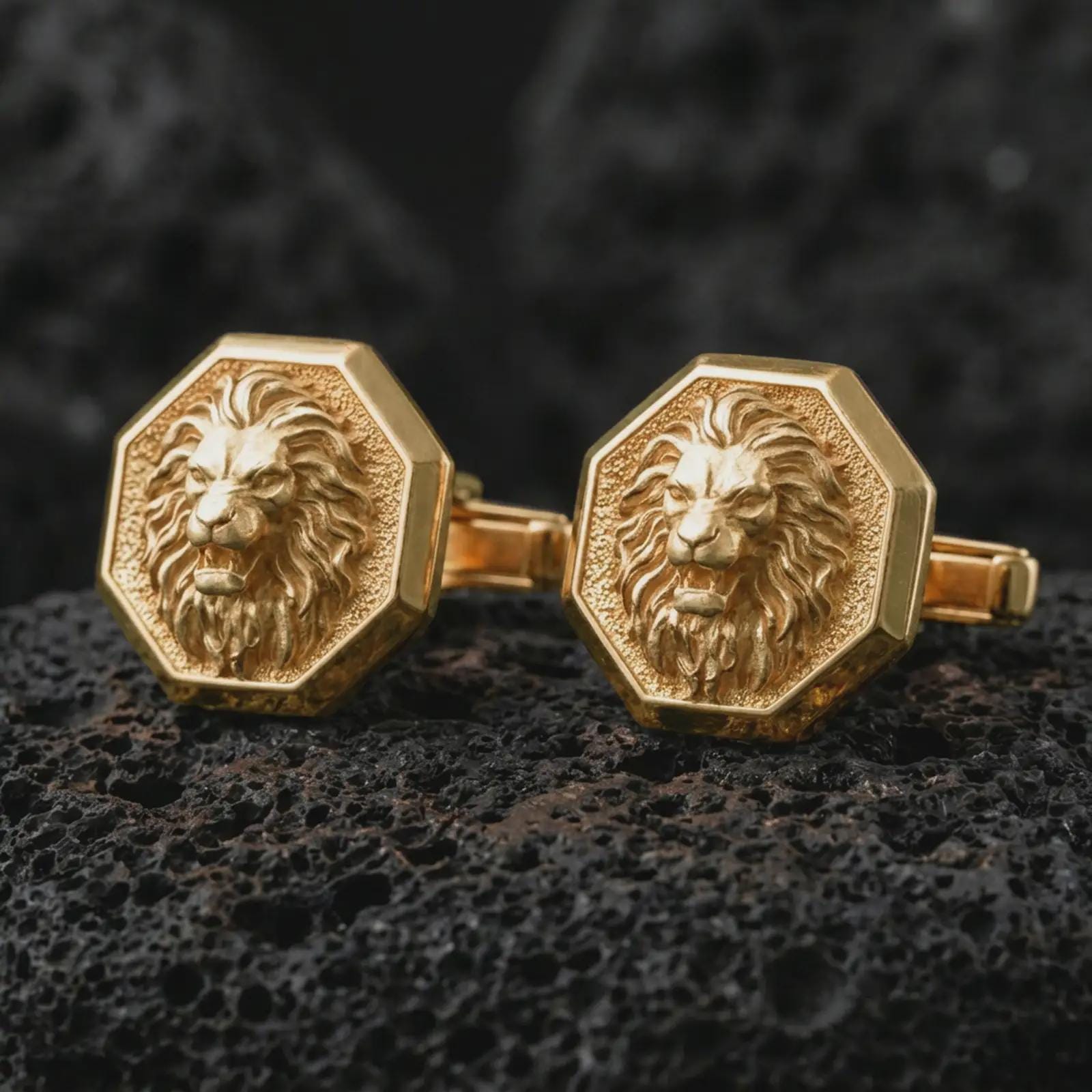 Gold plated lion head cufflinks sit on textured volcanic rock in a dramatic studio macro shot, highlighting the powerful animal motif and formal accessory design.
