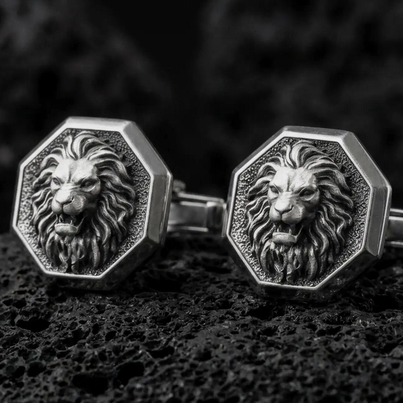 An oxidized lion head cufflink pair rests on dark volcanic rock in a macro studio close-up, showing a fierce roaring big cat with a sculpted mane inside octagon frames.