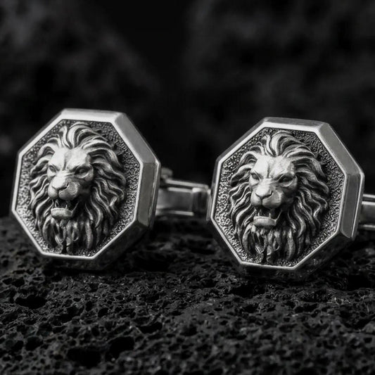 An oxidized lion head cufflink pair rests on dark volcanic rock in a macro studio close-up, showing a fierce roaring big cat with a sculpted mane inside octagon frames.