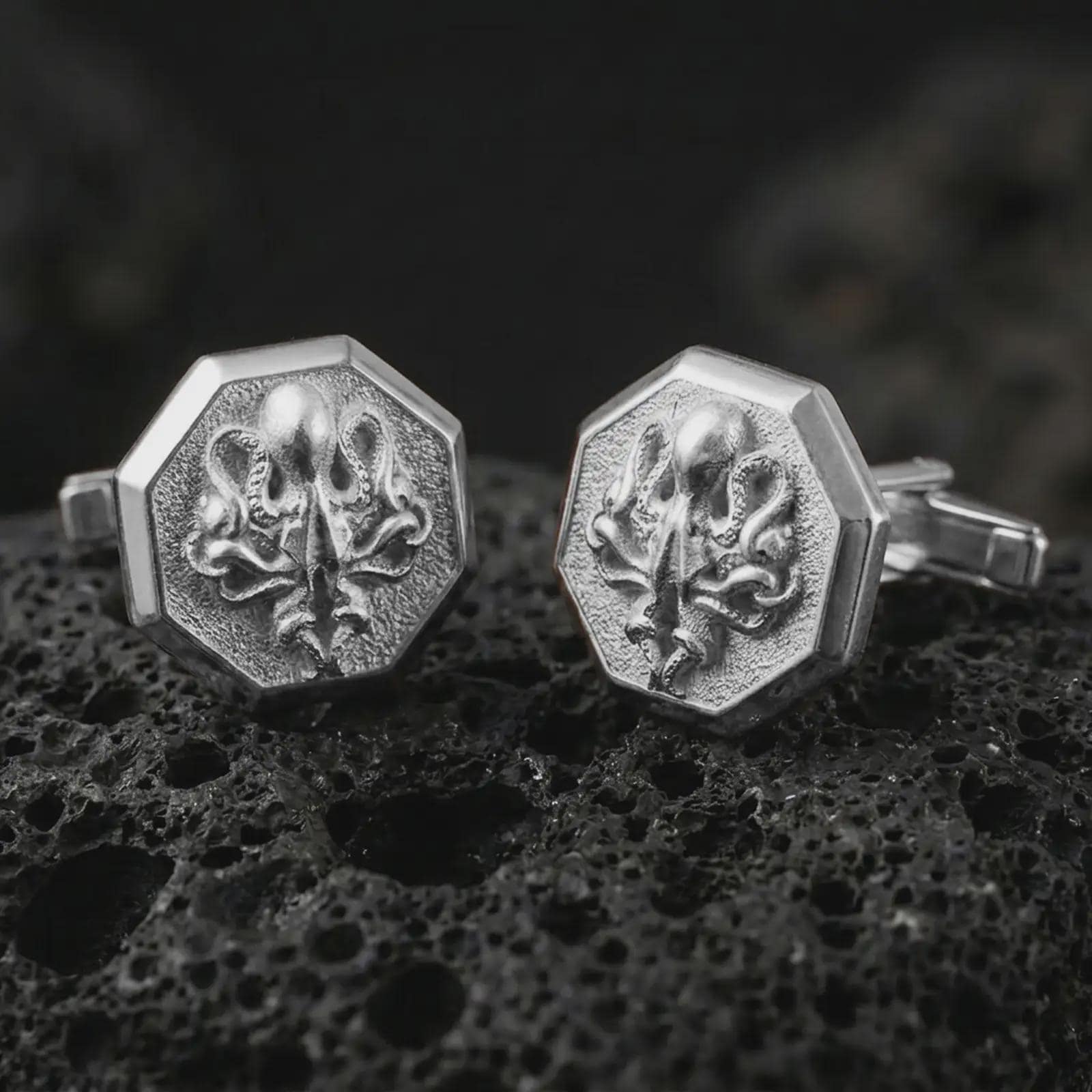 Polished silver octopus cufflinks are displayed on black volcanic rock in a premium macro studio close-up with detailed tentacle texture and clean geometric edges.