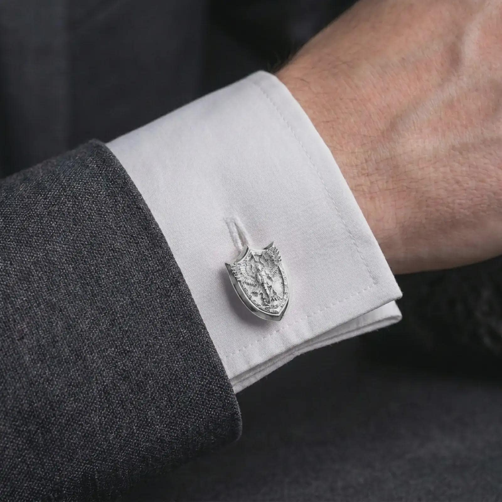A polished silver Saint Michael cufflink is worn on a shirt cuff in a premium close-up lifestyle image, showing the shield-shaped archangel relief clearly.