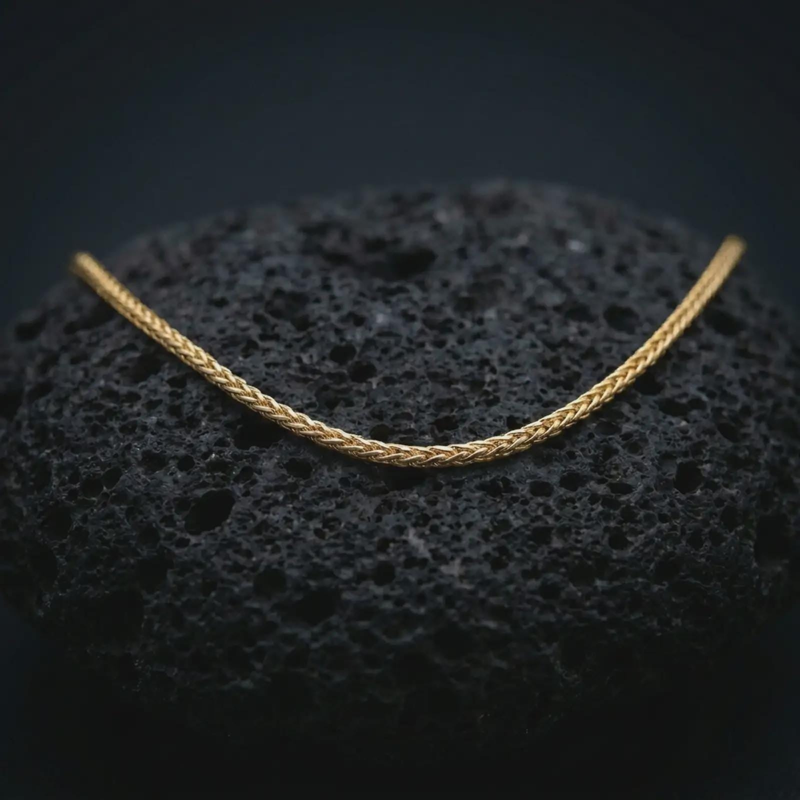 A gold plated foxtail chain necklace lies on textured volcanic rock in a dramatic studio macro shot, highlighting the 2.70mm woven chain structure.