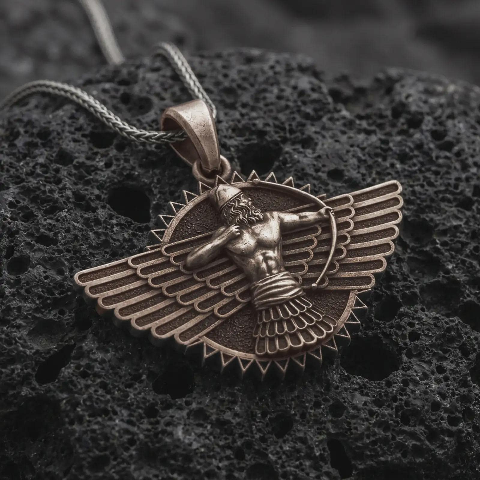 A bronze Faravahar pendant rests on dark volcanic rock in a macro studio close-up, showing a detailed winged Persian symbol with a central figure.