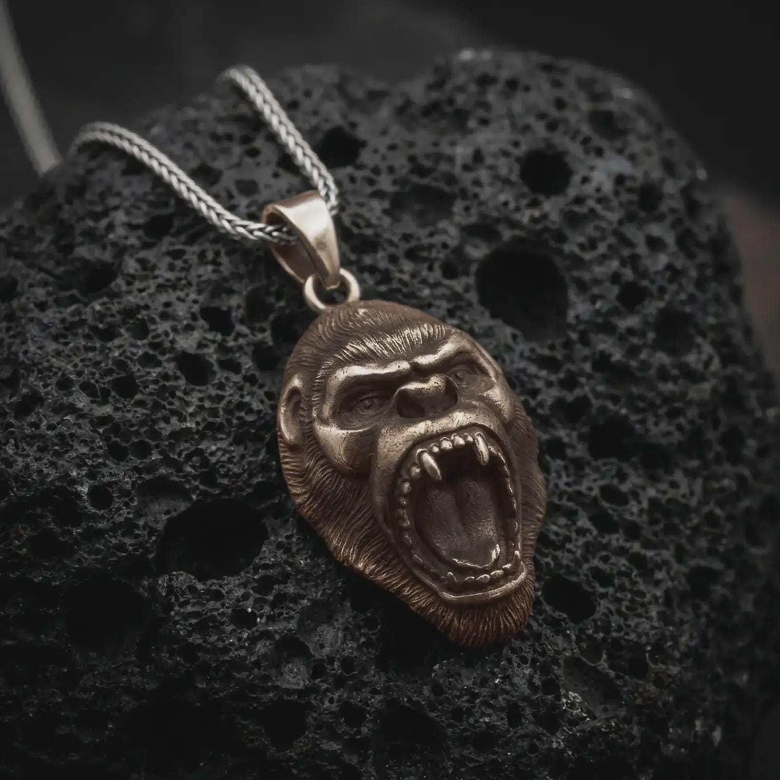 A bronze gorilla pendant rests on dark volcanic rock in a macro studio close-up, showing a fierce roaring ape head with detailed teeth and fur texture.
