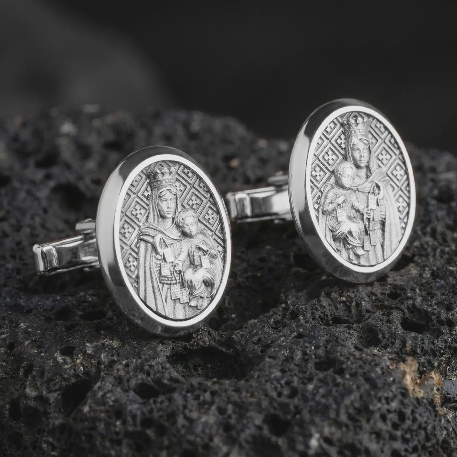 Polished silver Blessed Mother cufflinks are displayed on black volcanic rock in a premium macro studio close-up with detailed Catholic devotional artwork.