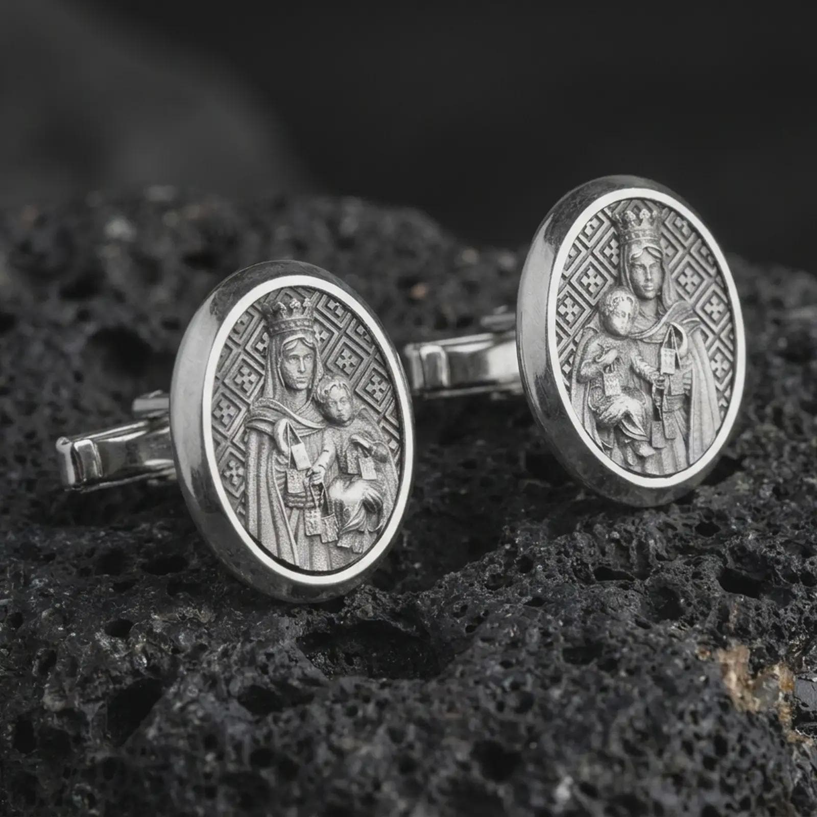 An oxidized Blessed Mother cufflink pair rests on dark volcanic rock in a macro studio close-up, showing the Virgin Mary and Child relief in oval frames.
