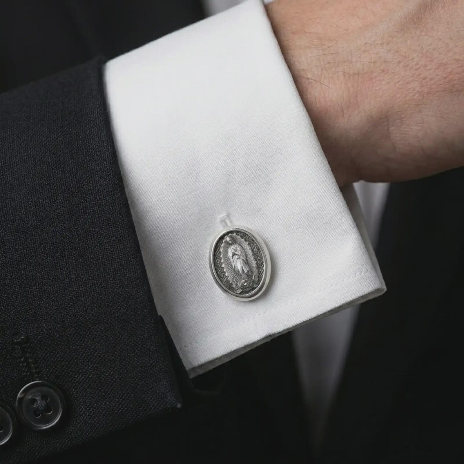 An oxidized Our Lady of Guadalupe cufflink is worn on a white shirt cuff in a close-up lifestyle shot, styled with a dark suit as a meaningful Catholic accessory.