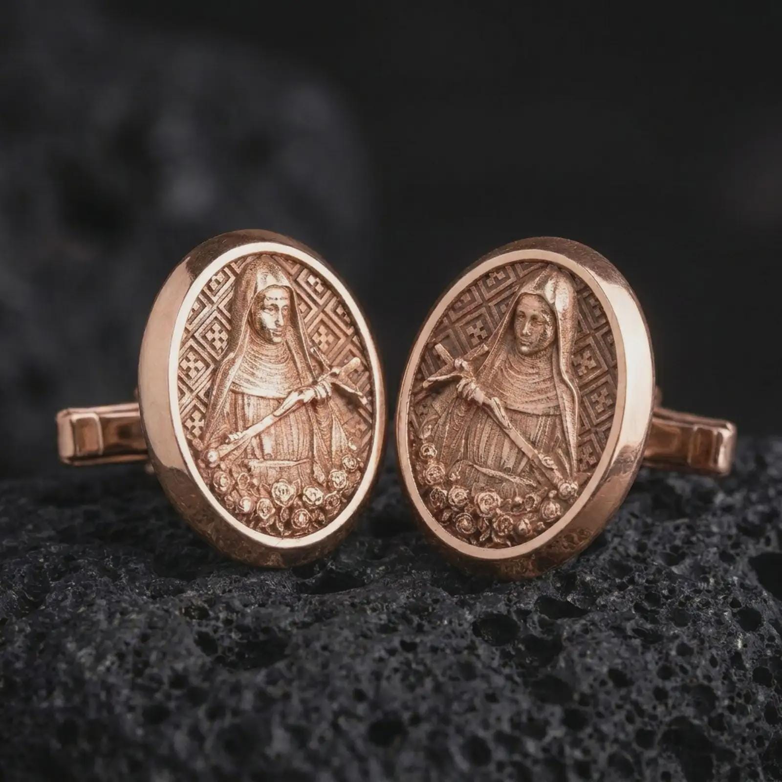 Rose gold plated Mary Magdalene cufflinks are photographed on porous volcanic rock in a close-up studio composition with refined luxury lighting and religious detail.