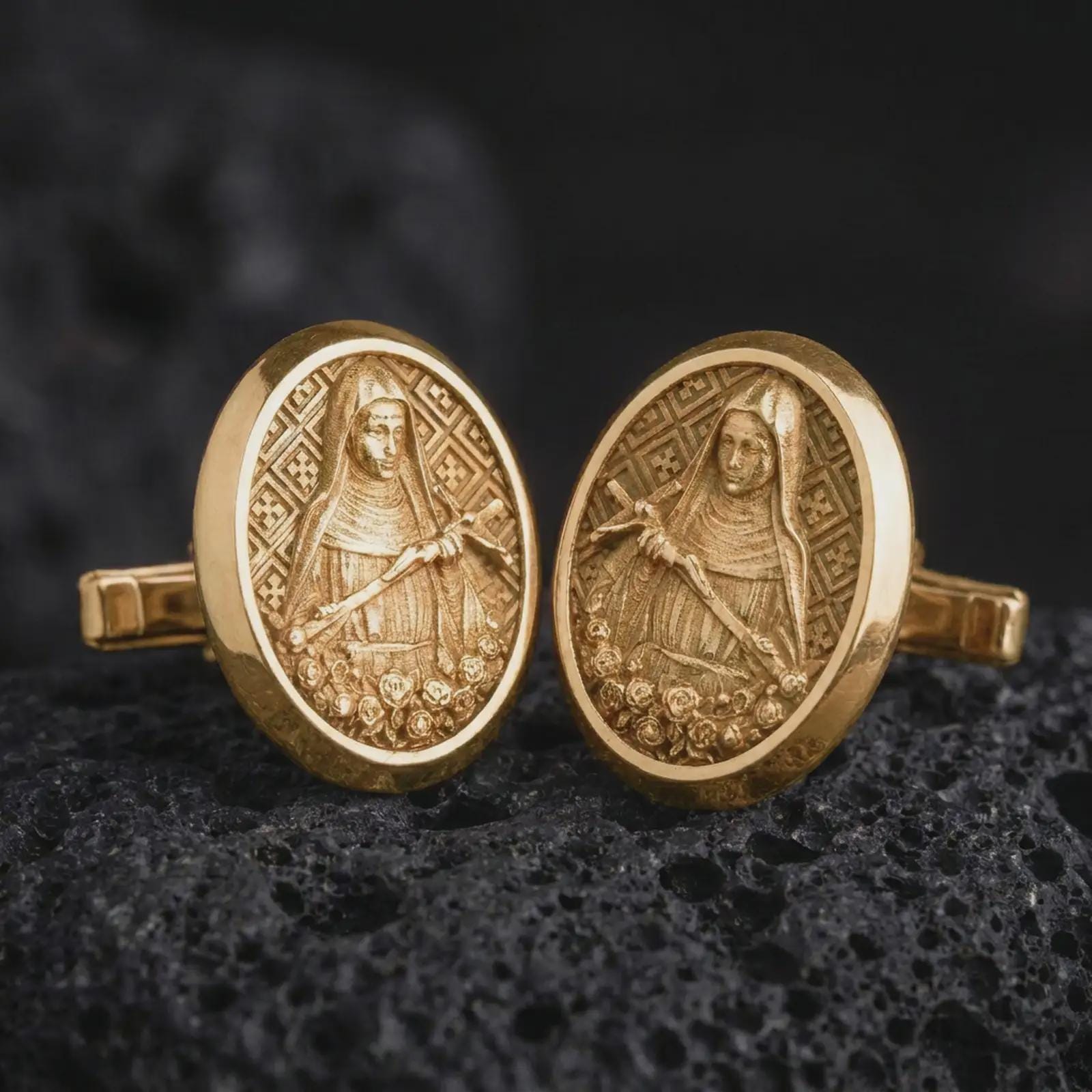 Gold plated Mary Magdalene cufflinks sit on textured volcanic rock in a dramatic studio macro shot, highlighting the sacred saint medallion design.
