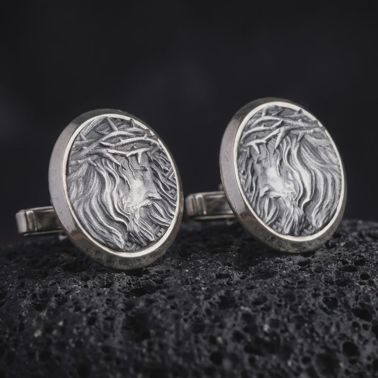 An oxidized Jesus Crown of Thorns cufflink pair rests on dark volcanic rock in a macro studio close-up, showing a detailed Christ portrait inside round frames.