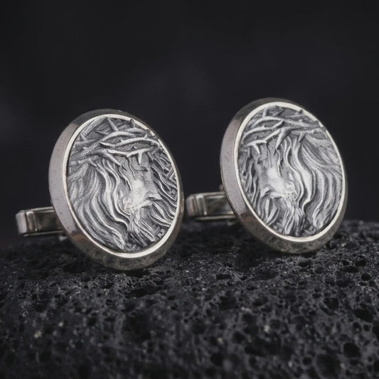 An oxidized Jesus Crown of Thorns cufflink pair rests on dark volcanic rock in a macro studio close-up, showing a detailed Christ portrait inside round frames.