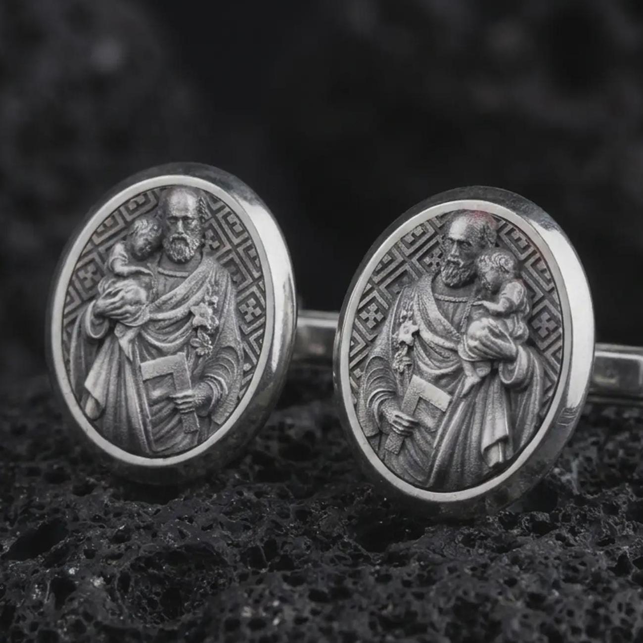 Oxidized Saint Joseph cufflinks displayed on dark volcanic rock in a macro studio photograph, featuring Saint Joseph holding the Christ Child with finely engraved Christian details.