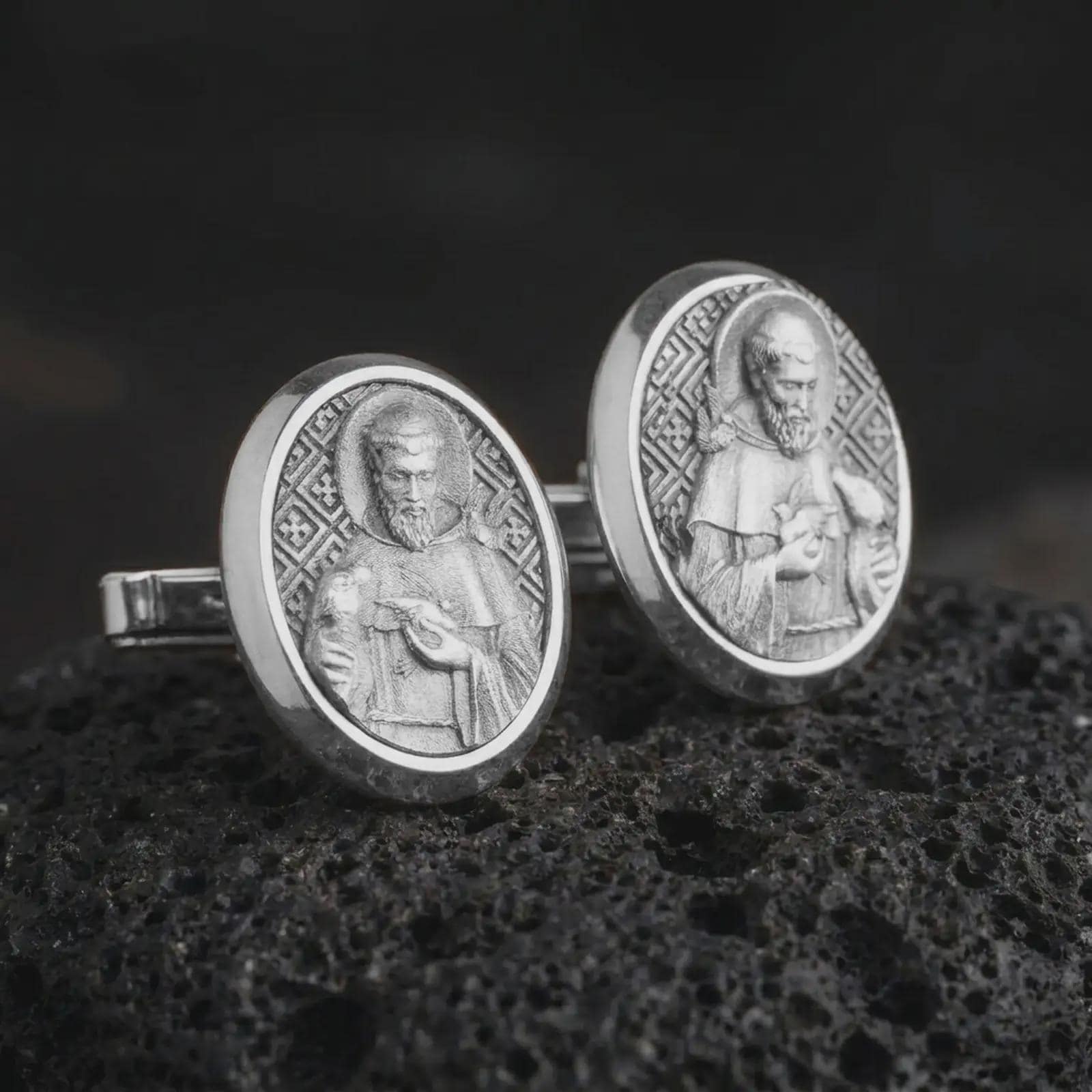 Polished silver Saint Francis cufflinks are displayed on black volcanic rock in a premium macro studio close-up with detailed Christian devotional artwork.