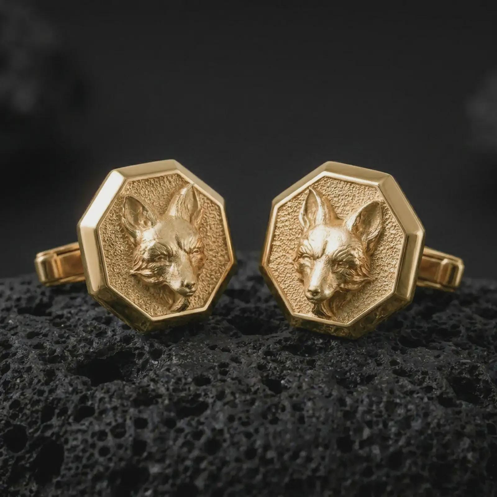 Gold plated fox head cufflinks sit on textured volcanic rock in a dramatic studio macro shot, highlighting the clever animal motif and formal accessory design.