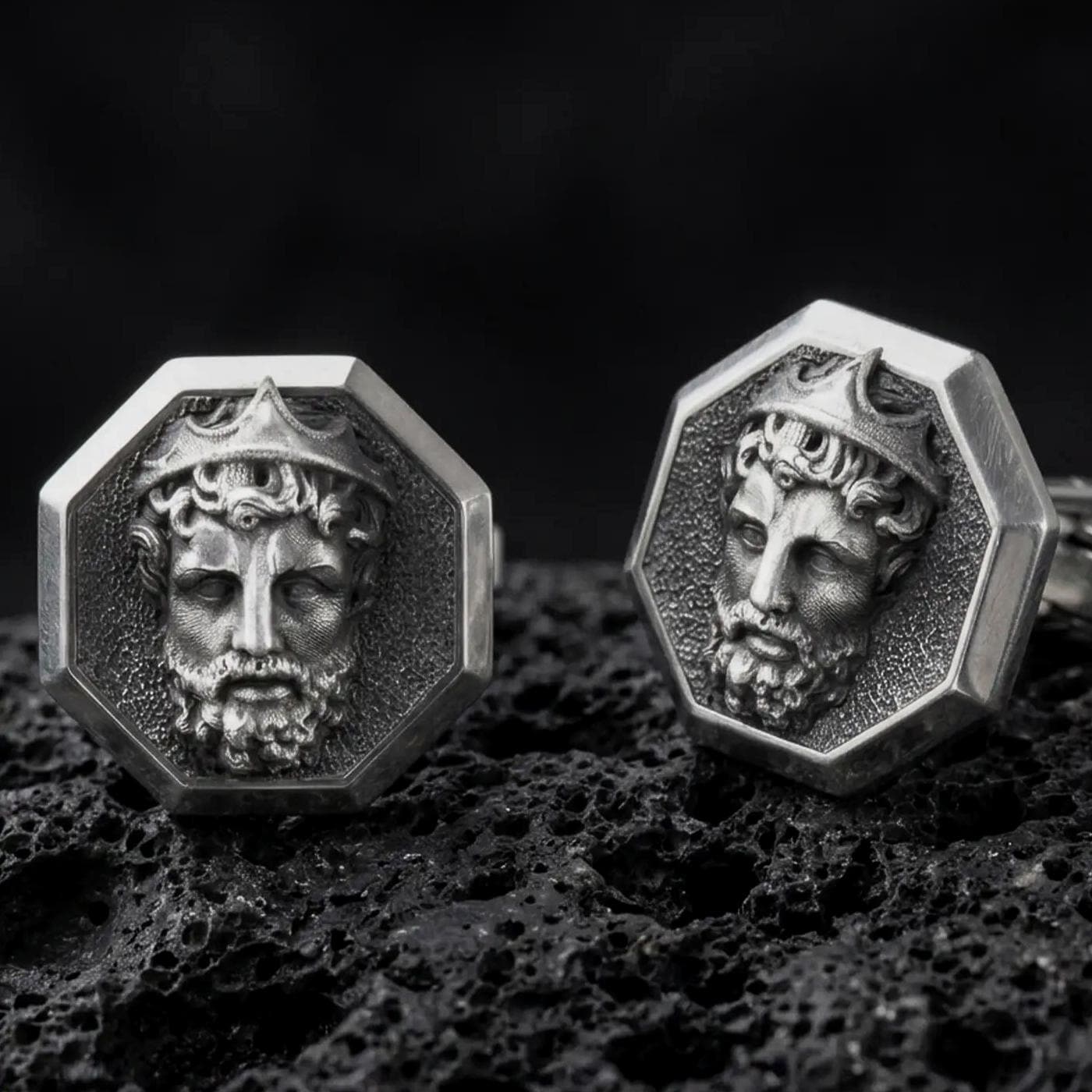 An oxidized Zeus cufflink pair rests on dark volcanic rock in a macro studio close-up, showing a sculpted Greek god portrait inside octagon frames.