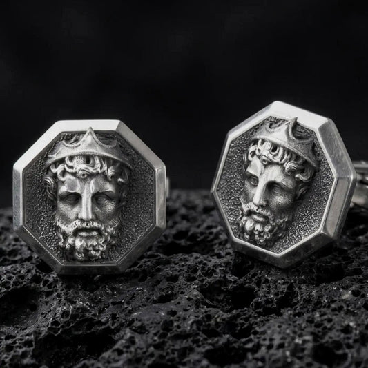 An oxidized Zeus cufflink pair rests on dark volcanic rock in a macro studio close-up, showing a sculpted Greek god portrait inside octagon frames.