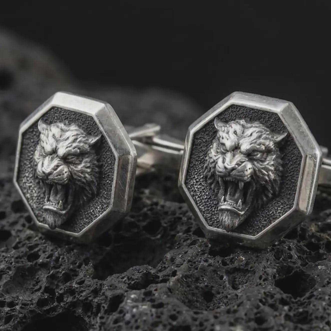 An oxidized tiger head cufflink pair rests on dark volcanic rock in a macro studio close-up, showing a fierce roaring big cat inside octagon frames.