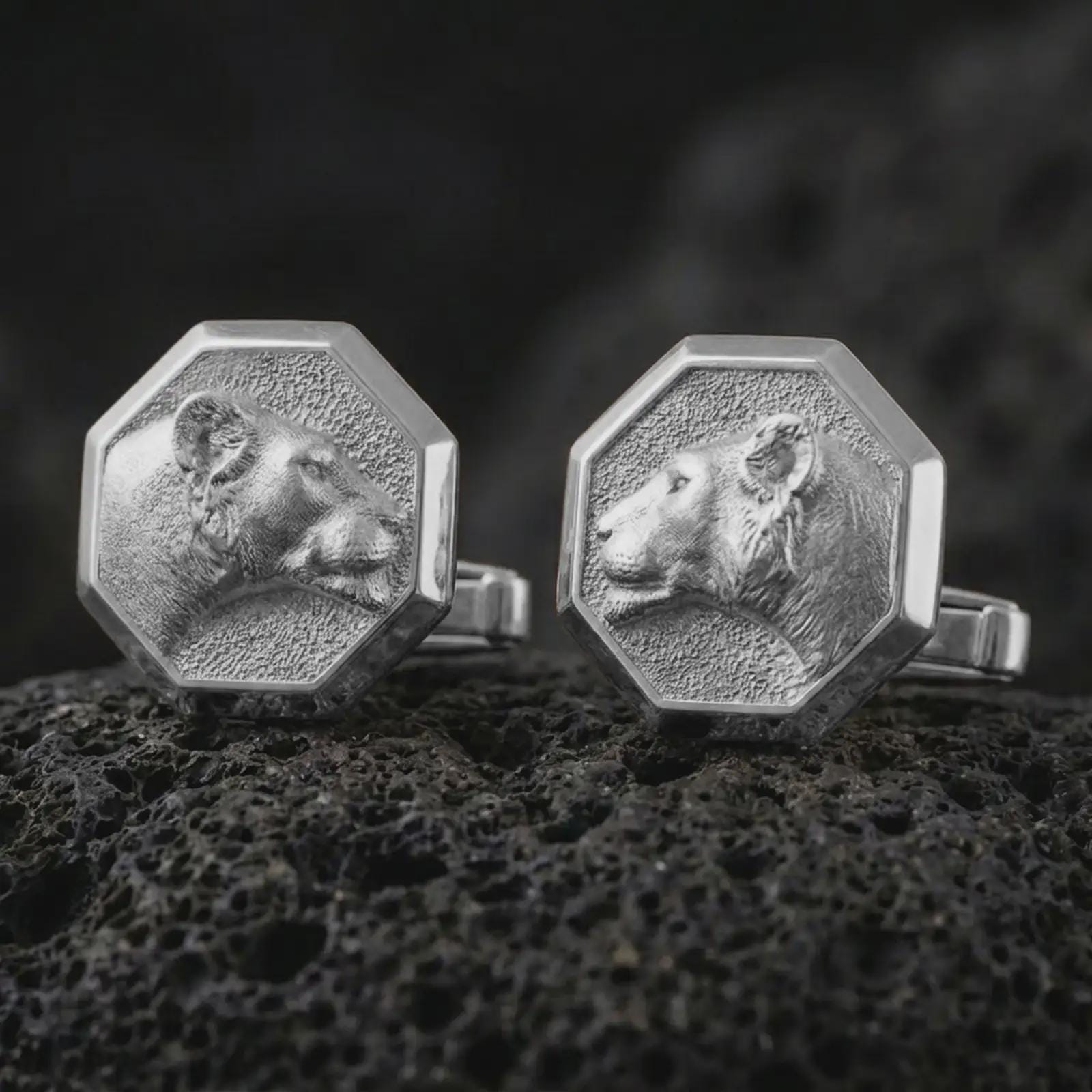 Polished silver lioness head cufflinks are displayed on black volcanic rock in a premium macro studio close-up with soft facial texture and clean geometric edges.