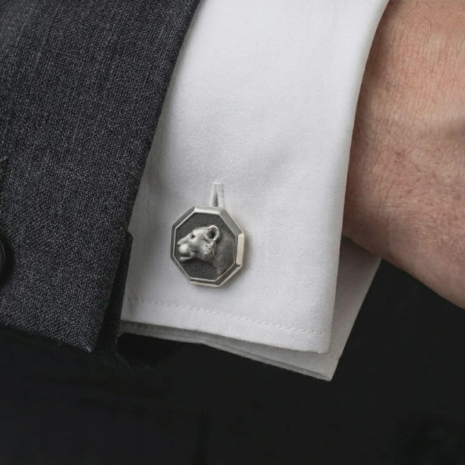 An oxidized lioness head cufflink is worn on a white shirt cuff in a close-up lifestyle shot, styled with a dark suit for a refined wildlife-inspired look.