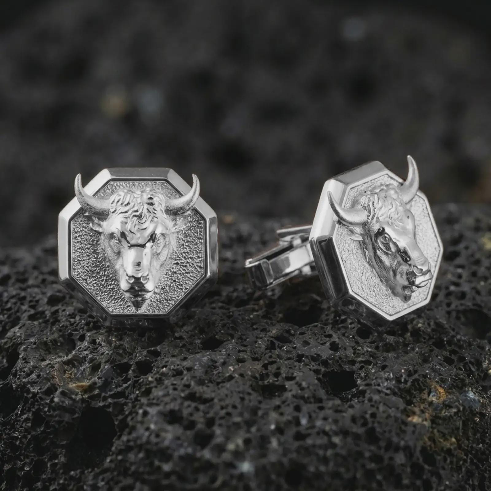 Polished silver bull head cufflinks are displayed on black volcanic rock in a premium macro studio close-up with detailed horns and clean geometric edges.