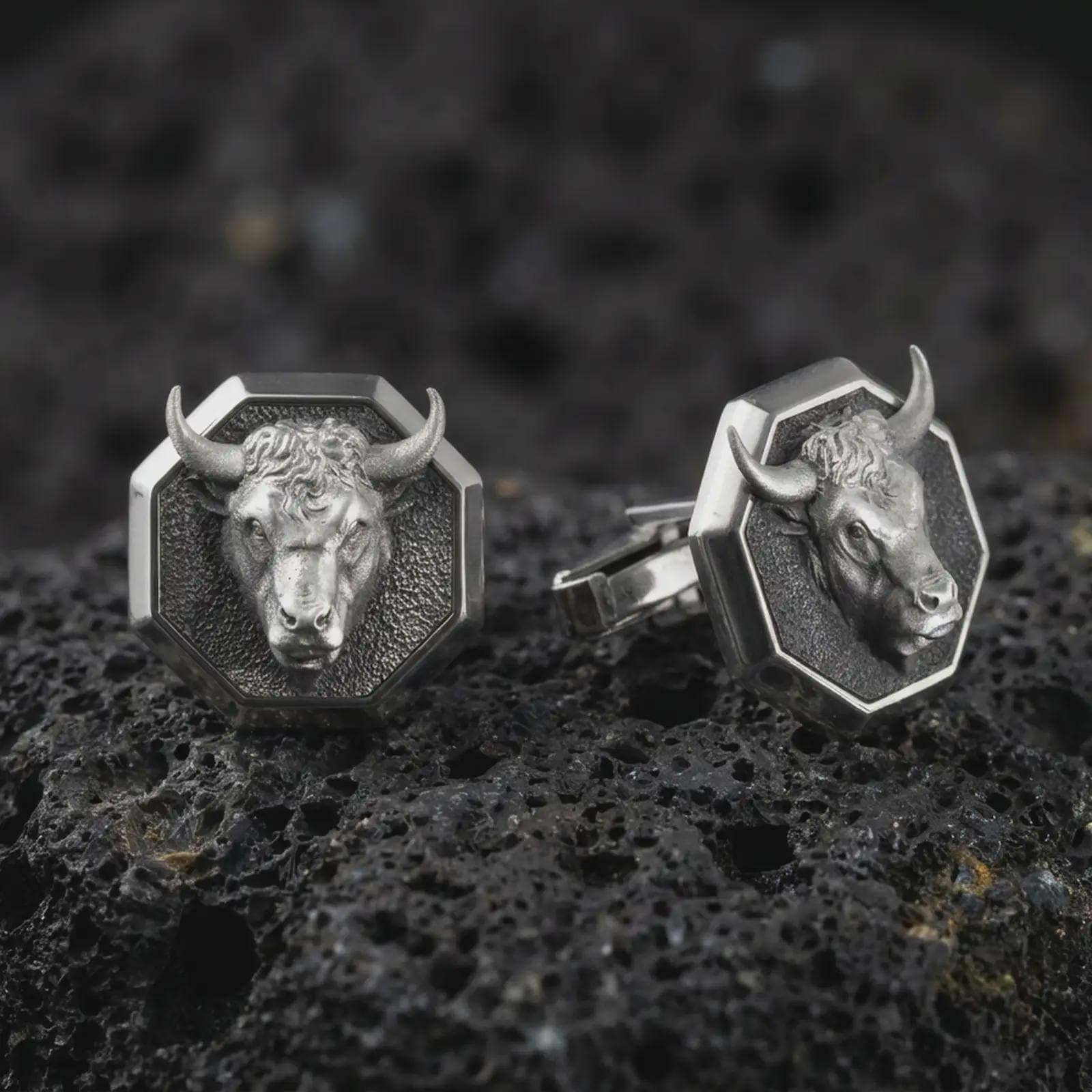 An oxidized bull head cufflink pair rests on dark volcanic rock in a macro studio close-up, showing a sculpted horned animal face inside octagon frames.