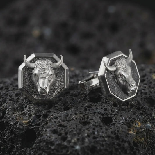 An oxidized bull head cufflink pair rests on dark volcanic rock in a macro studio close-up, showing a sculpted horned animal face inside octagon frames.