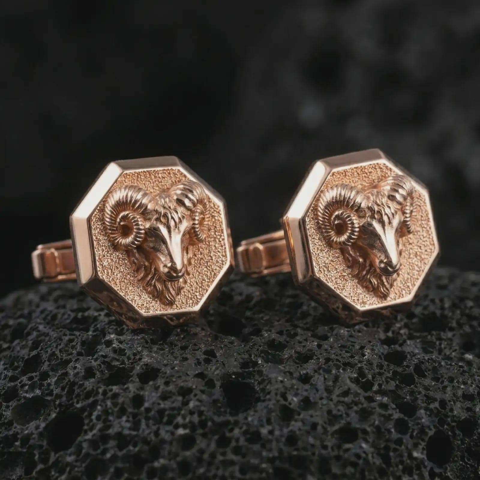Rose gold plated ram head cufflinks are photographed on porous volcanic rock in a close-up studio composition with refined luxury lighting and strong ram features.
