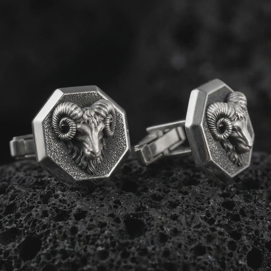 An oxidized ram head cufflink pair rests on dark volcanic rock in a macro studio close-up, showing sculpted curled horns and animal detail inside octagon frames.