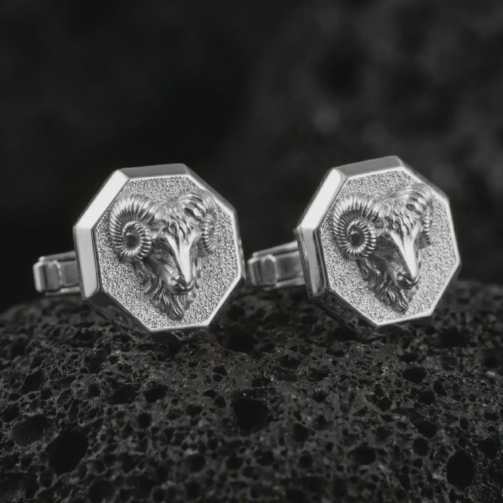 Polished silver ram head cufflinks are displayed on black volcanic rock in a premium macro studio close-up with detailed horn texture and clean geometric edges.