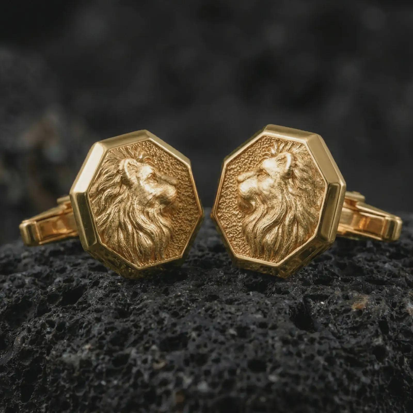 Gold plated lion profile cufflinks sit on textured volcanic rock in a dramatic studio macro shot, highlighting the noble animal motif and formal accessory design.