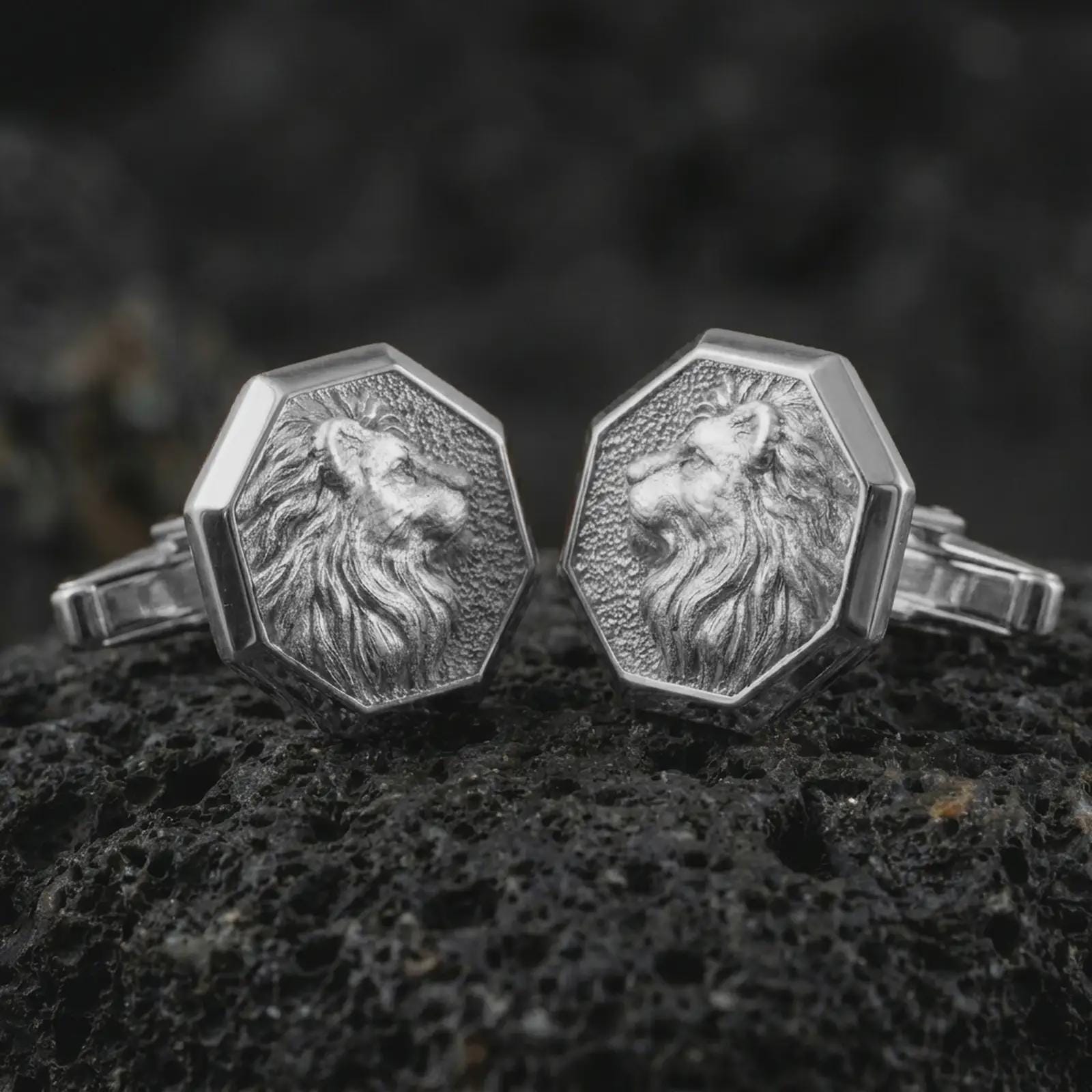 Polished silver lion profile cufflinks are displayed on black volcanic rock in a premium macro studio close-up with detailed mane texture and clean geometric edges.