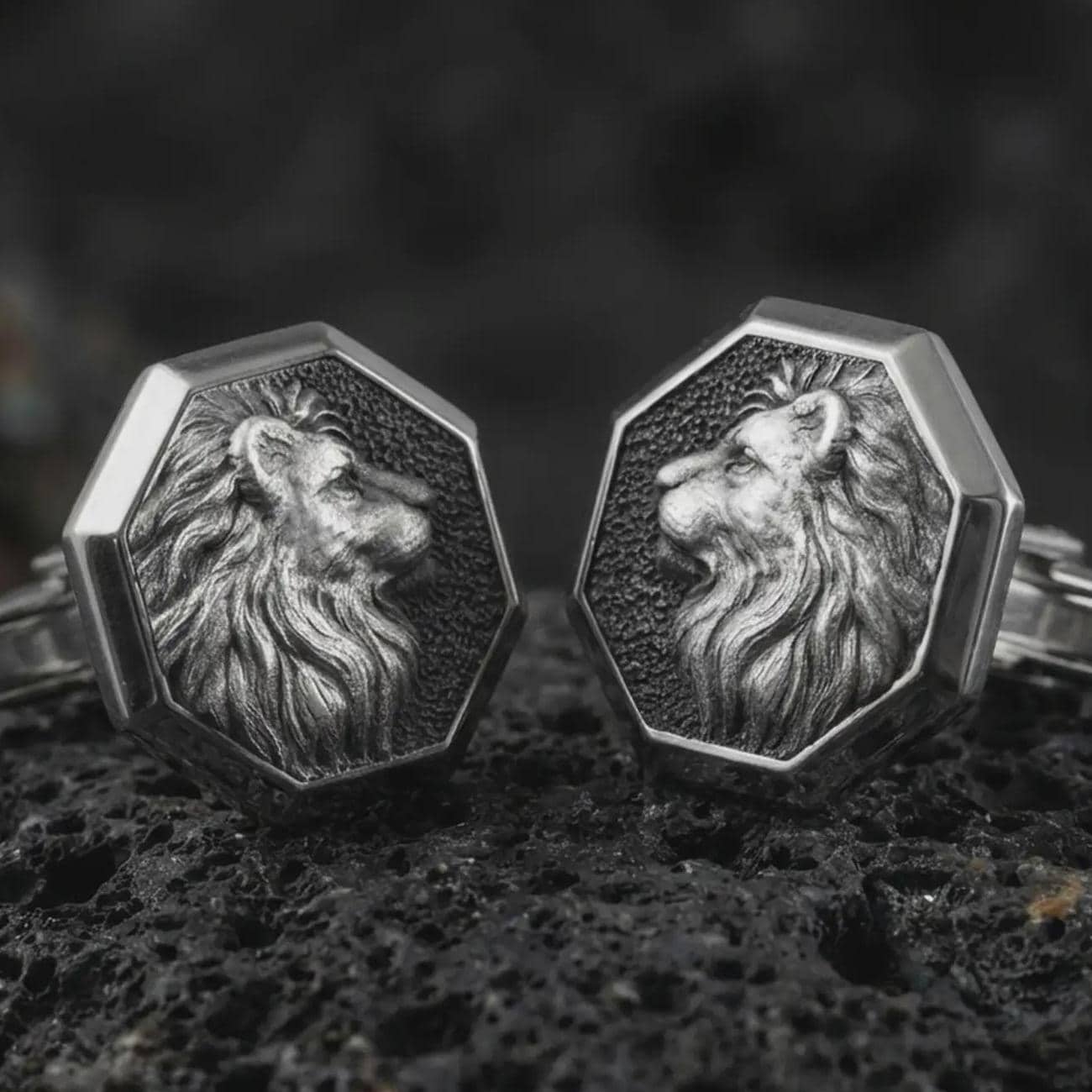 An oxidized lion profile cufflink pair rests on dark volcanic rock in a macro studio close-up, showing a sculpted big cat head with flowing mane inside octagon frames.
