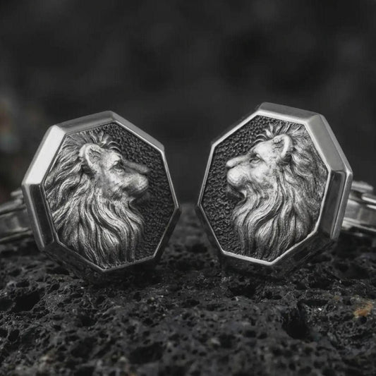 An oxidized lion profile cufflink pair rests on dark volcanic rock in a macro studio close-up, showing a sculpted big cat head with flowing mane inside octagon frames.