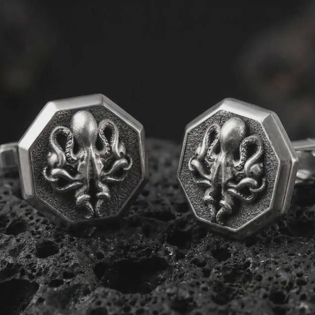 An oxidized octopus cufflink pair rests on dark volcanic rock in a macro studio close-up, showing sculpted tentacles and sea creature detail inside octagon frames.