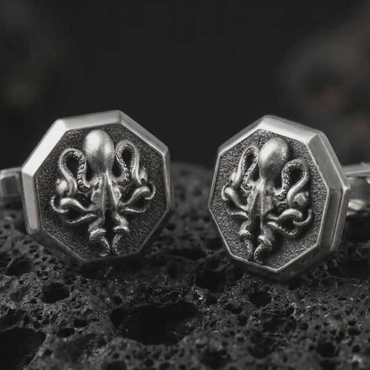 An oxidized octopus cufflink pair rests on dark volcanic rock in a macro studio close-up, showing sculpted tentacles and sea creature detail inside octagon frames.