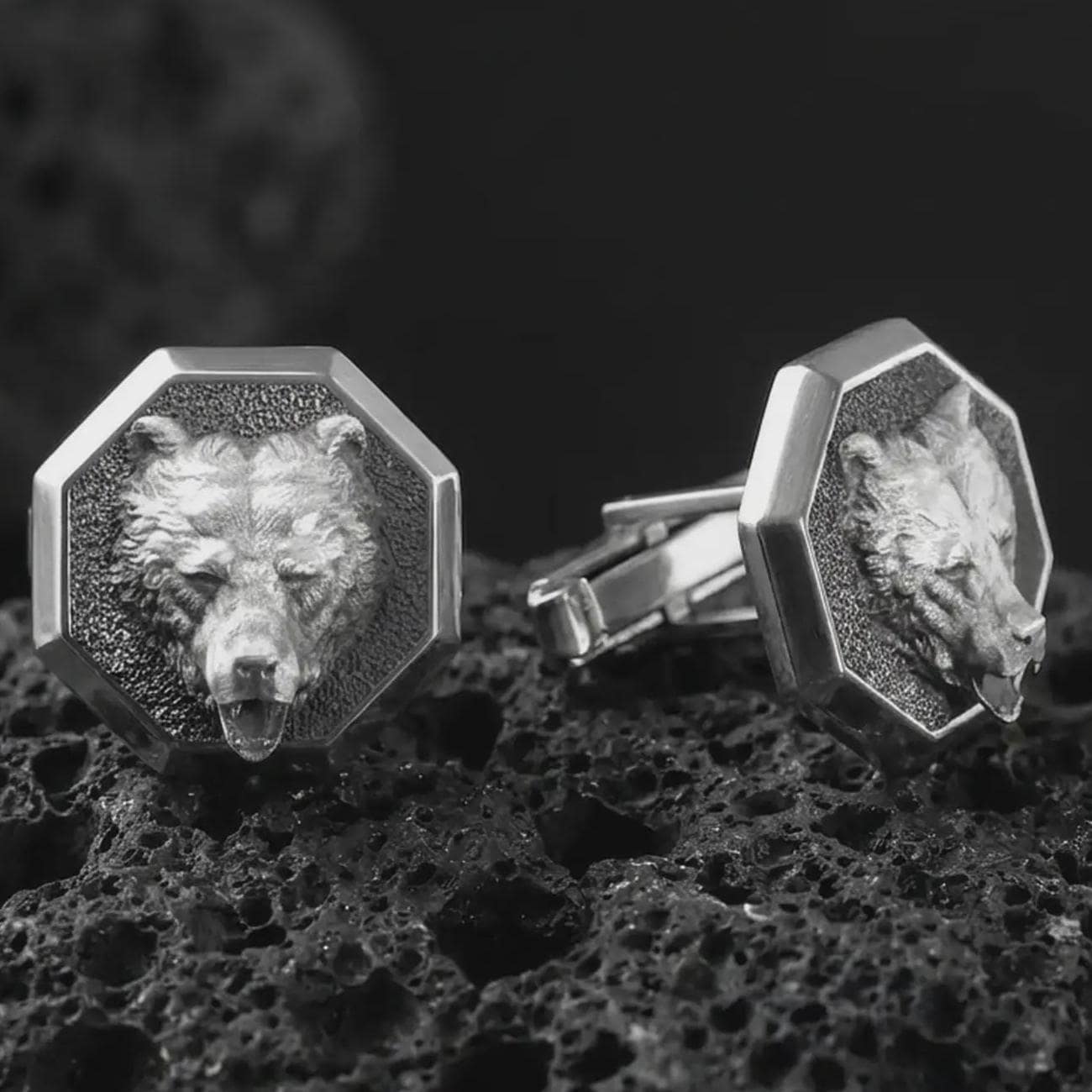 An oxidized bear head cufflink pair rests on dark volcanic rock in a macro studio close-up, showing a sculpted grizzly-inspired animal face inside octagon frames.